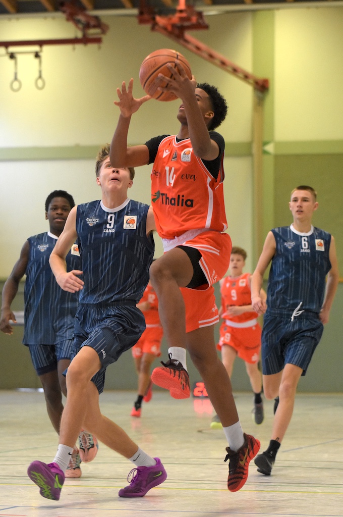 Ein junger Basketballspieler im orangefarbenen Trikot springt mit konzentriertem Gesichtsausdruck zum Korbleger, während Gegner in blauen Trikots hinter ihm herlaufen