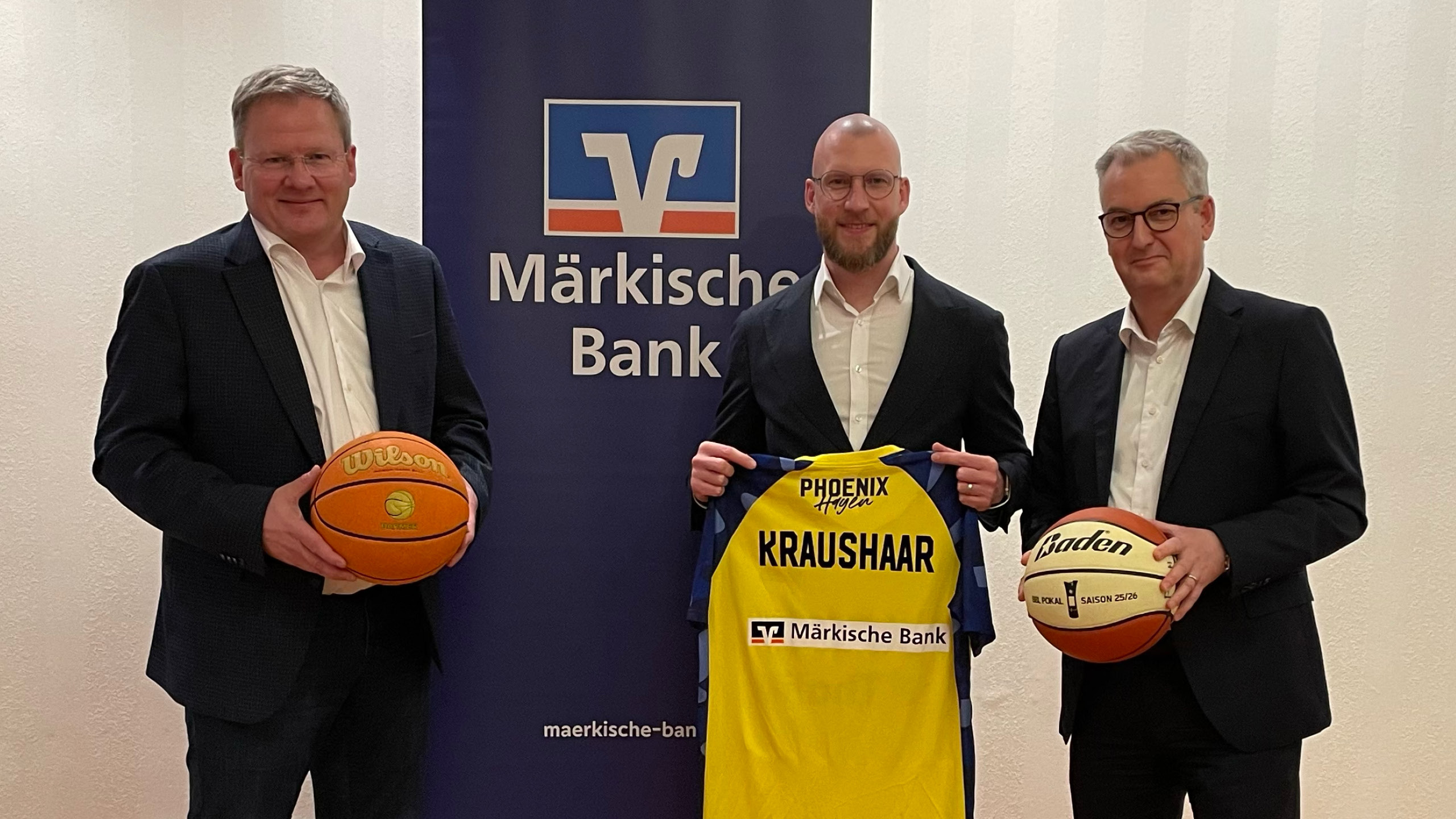 Drei Männer in Anzügen posieren freundlich mit Basketbällen und gelbem Trikot
