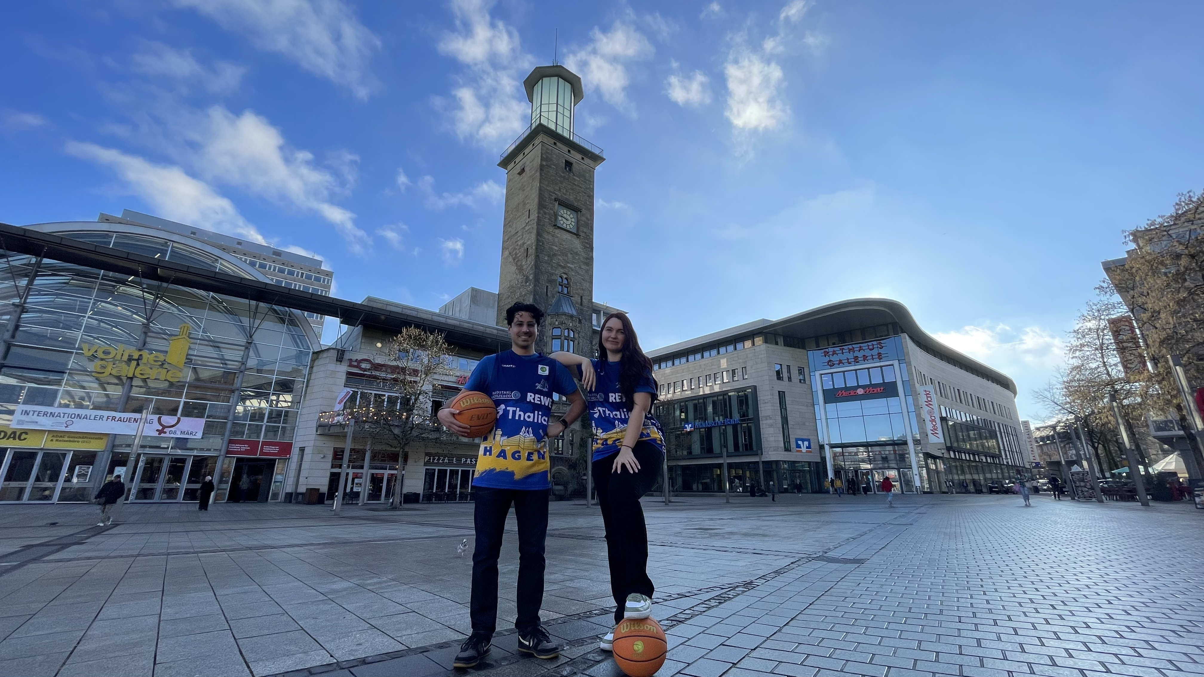 Zwei junge Menschen in blauen Basketballtrikots stehen lächelnd mit Bällen auf einem Platz