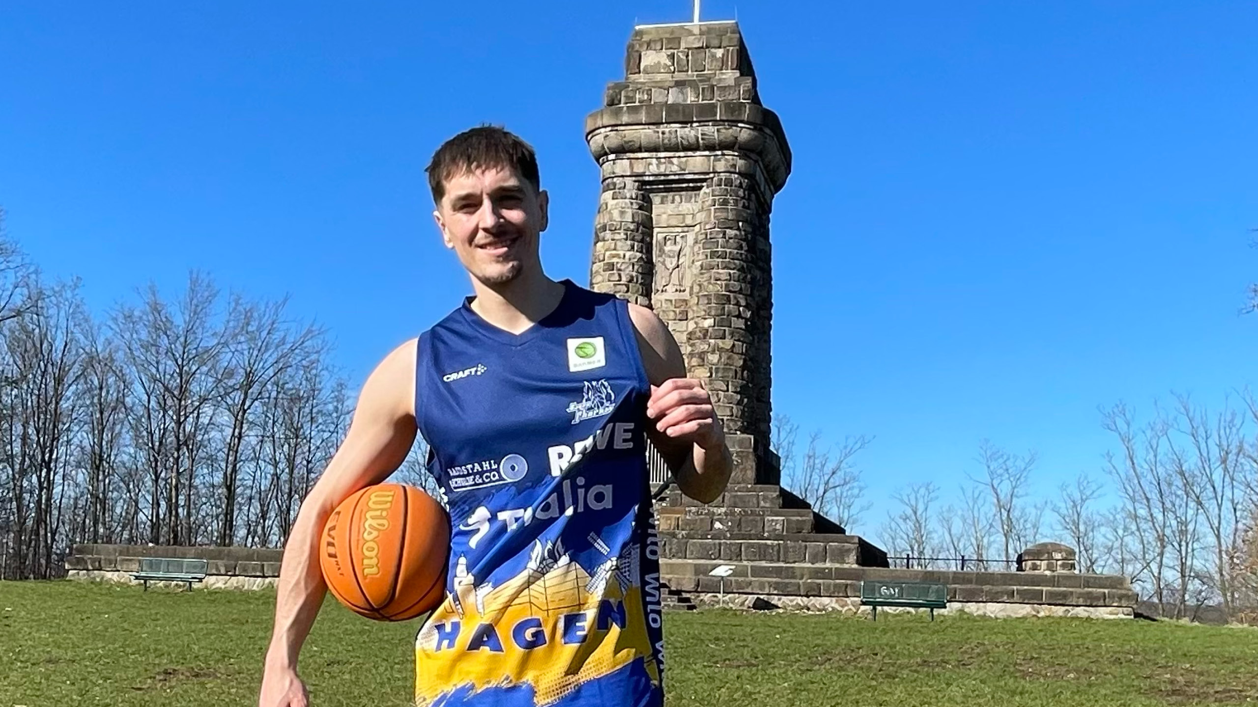 Ein junger Mann mit sportlichem Gesichtsausdruck hält einen Basketball vor einem alten Steinturm in der Sonne