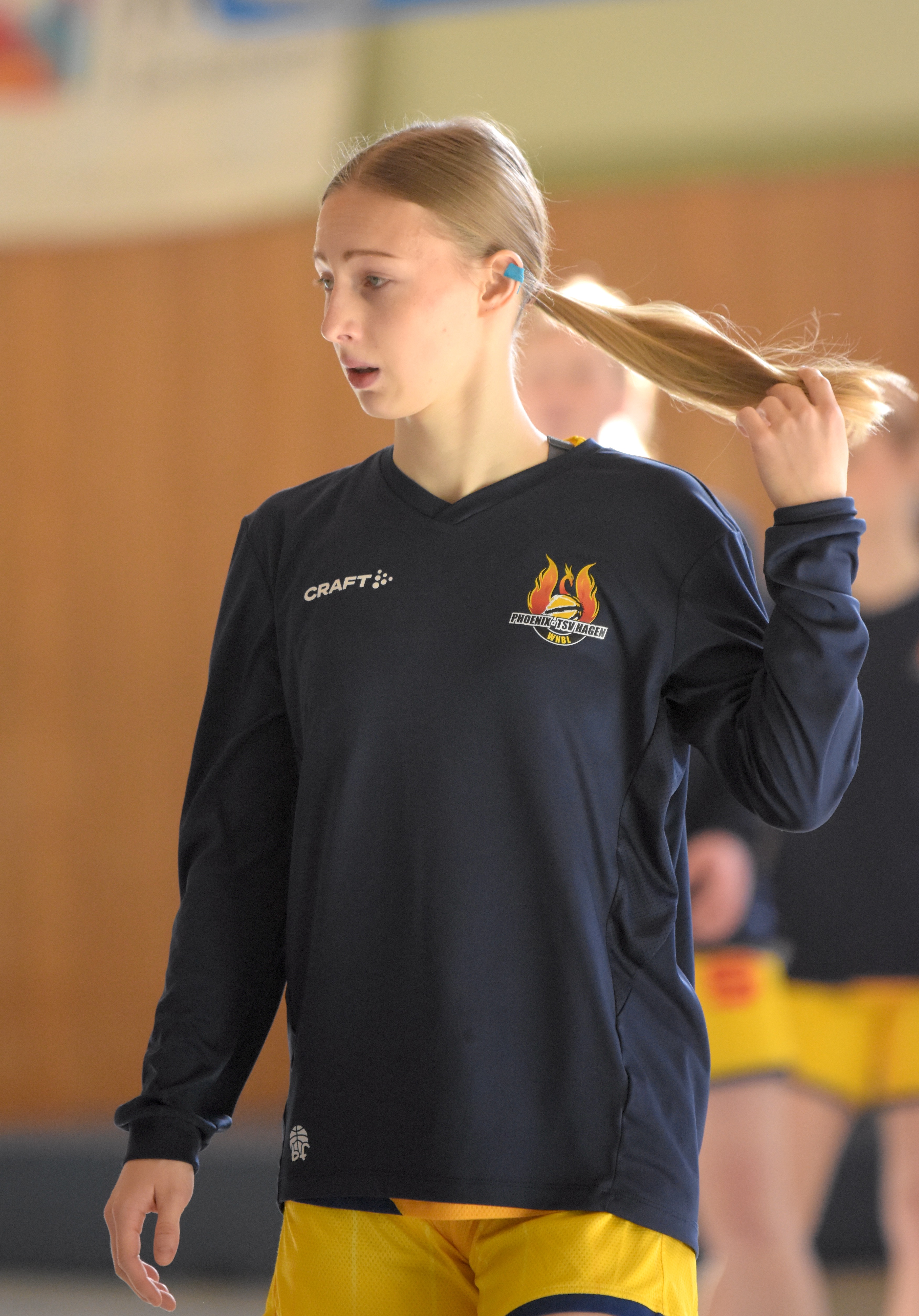 Eine junge Frau mit blondem Zopf und neutralem Gesichtsausdruck trägt ein dunkelblaues Oberteil und hält ihre Haare