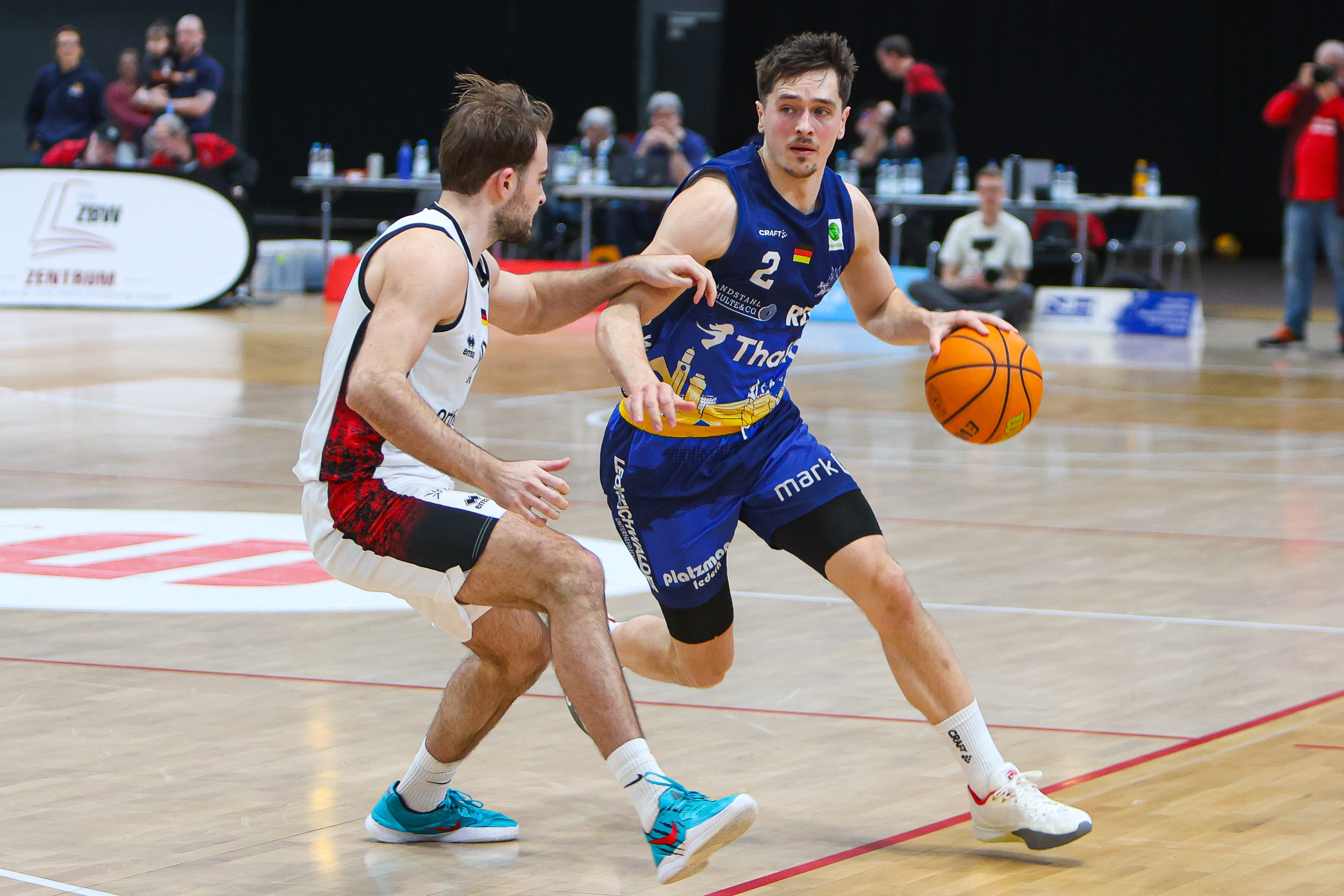 Zwei Basketballspieler im Spiel, einer im blauen Trikot mit entschlossenem Blick und Ball