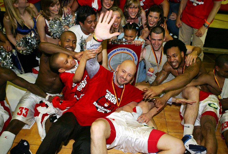 Mehrere jubelnde Basketballspieler und Cheerleader posieren lachend mit einem großen Pokal