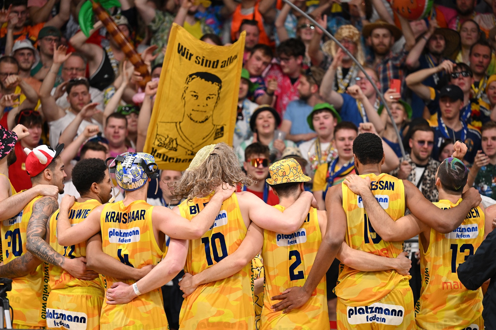 Eine Gruppe jubelnder Basketballspieler im gelben Trikot feiert Arm in Arm mit Fans