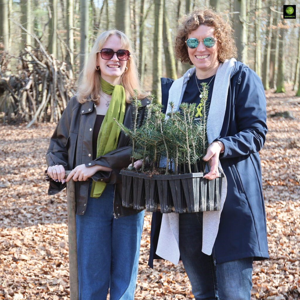 Zwei lächelnde Frauen mit Sonnenbrillen halten im sonnigen Wald junge grüne Pflanzen in schwarzen Behältern