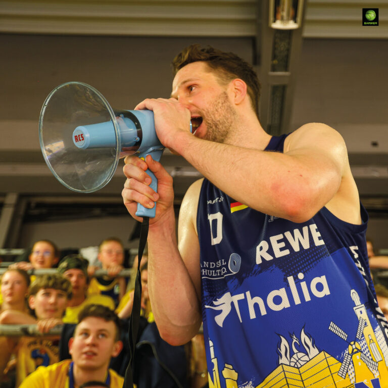 Ein Mann im blauen Basketballtrikot ruft mit einem Megafon fröhlich in die Menge