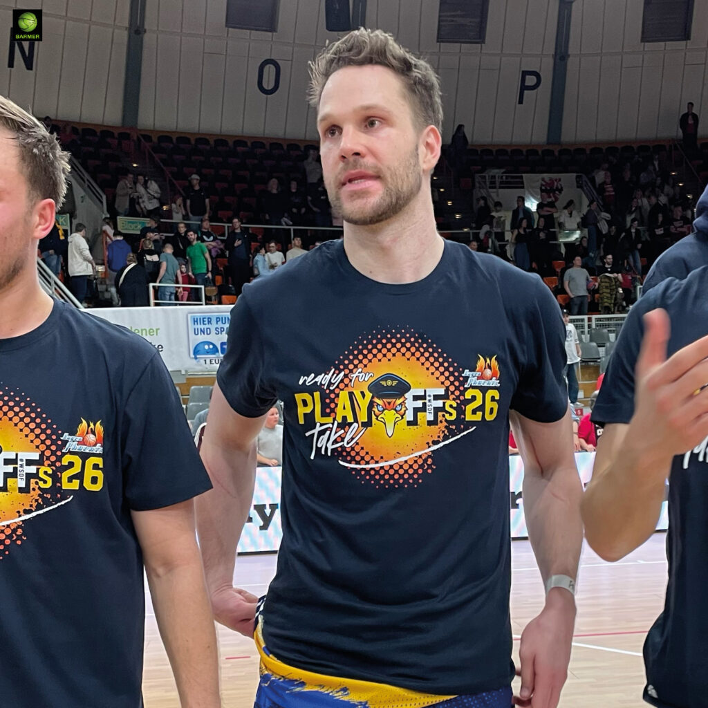Ein großer Mann mit kurzem braunem Haar und entspanntem Gesichtsausdruck steht in einer Sporthalle und trägt ein schwarzes Playoffs 26 T Shirt