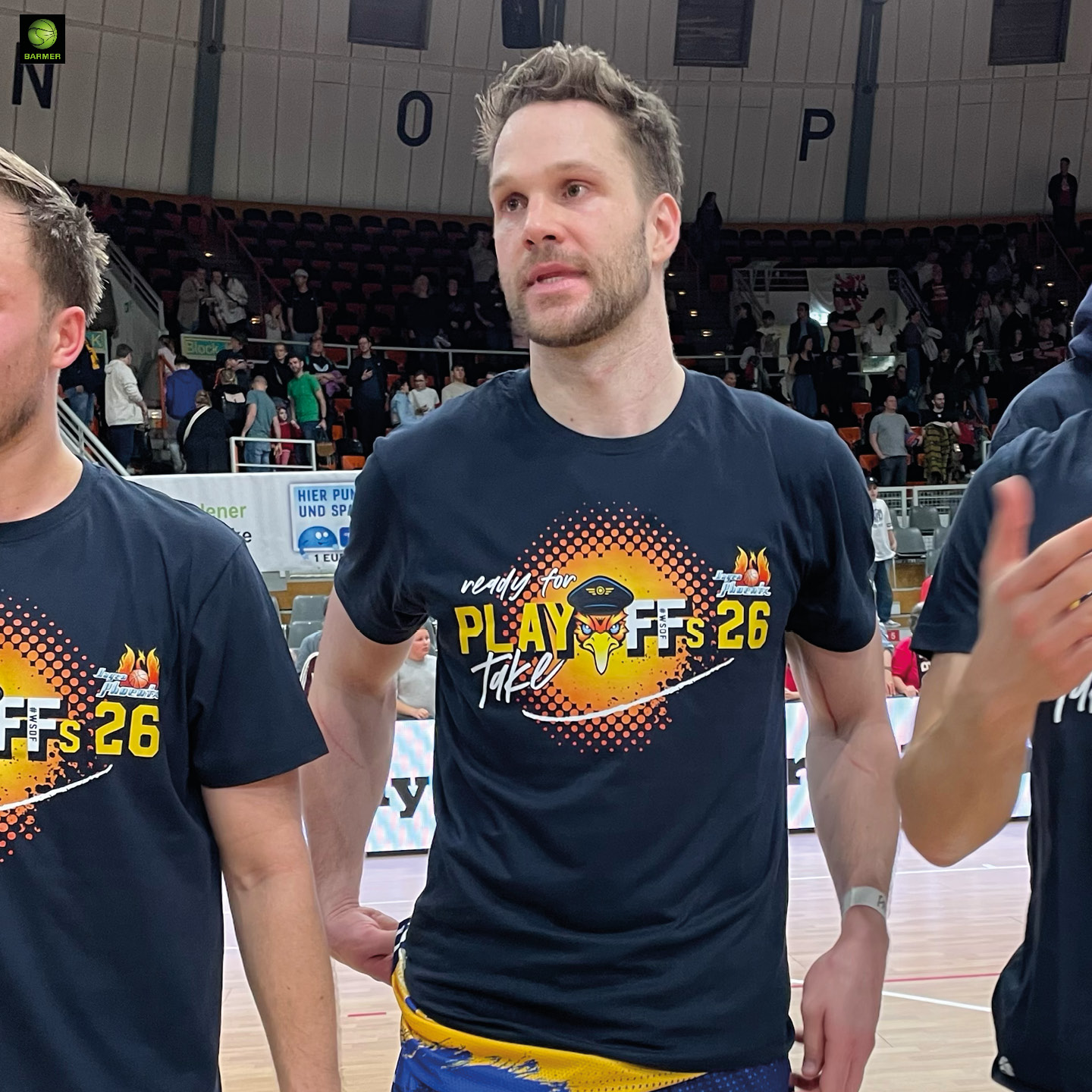 Ein großer Mann mit kurzem braunem Haar und entspanntem Gesichtsausdruck steht in einer Sporthalle und trägt ein schwarzes Playoffs 26 T Shirt