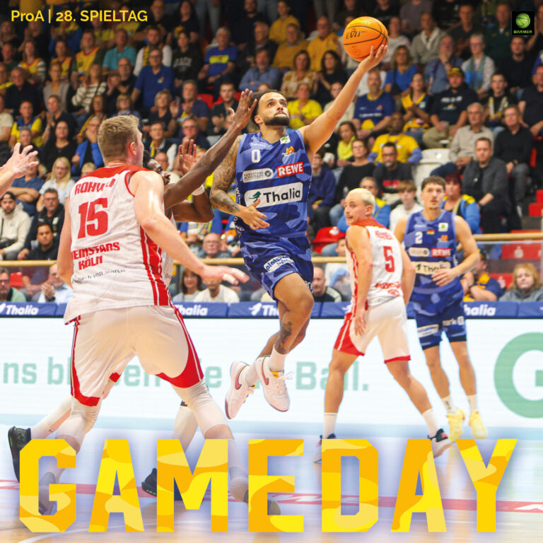 Ein Basketballspieler im blauen Trikot springt entschlossen mit dem Ball zum Korb, während Gegner in weißen Trikots ihn umgeben