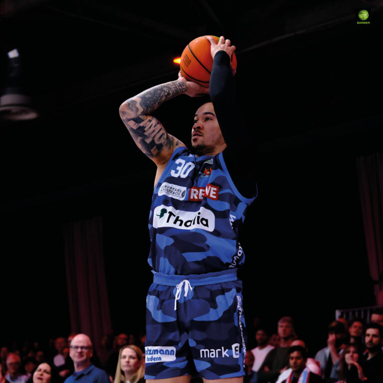 Ein tätowierter Basketballspieler im blauen Trikot konzentriert sich beim Wurf