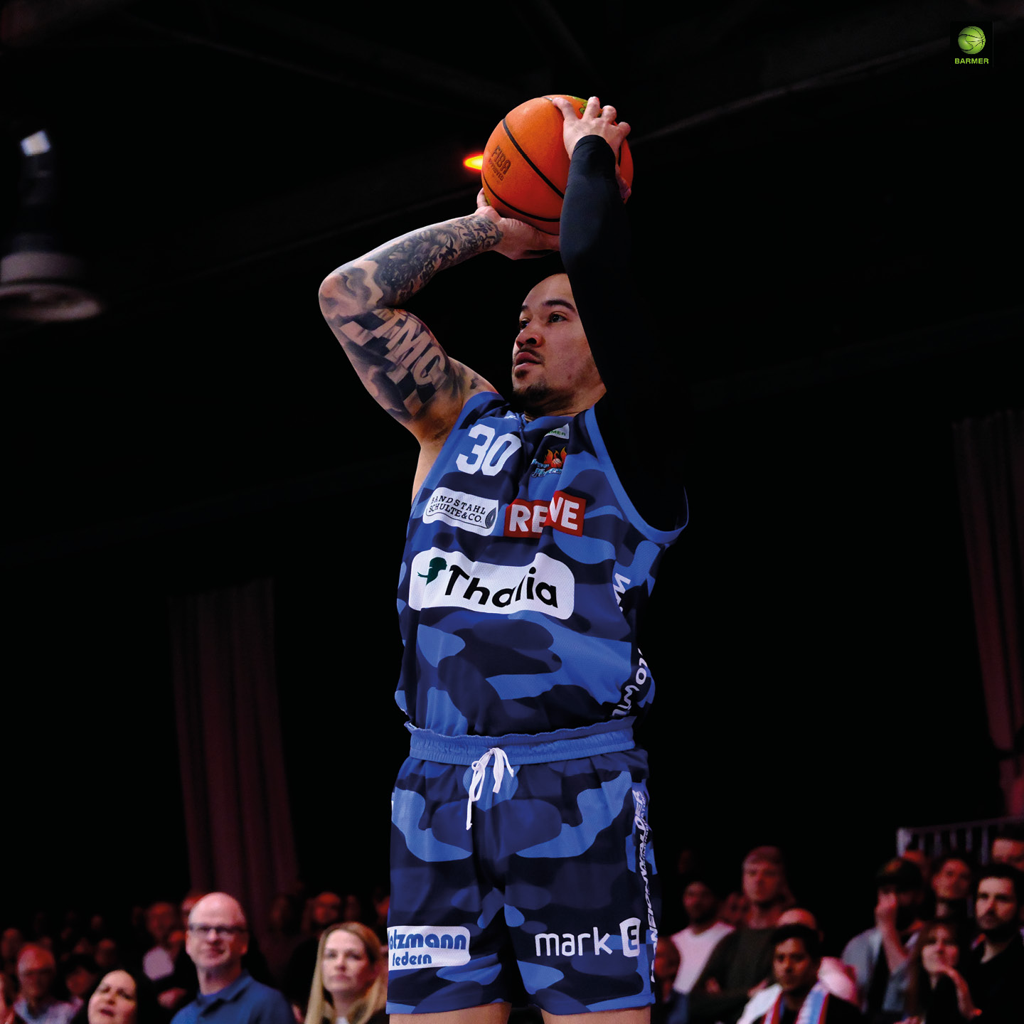 Ein tätowierter Basketballspieler im blauen Trikot konzentriert sich beim Wurf