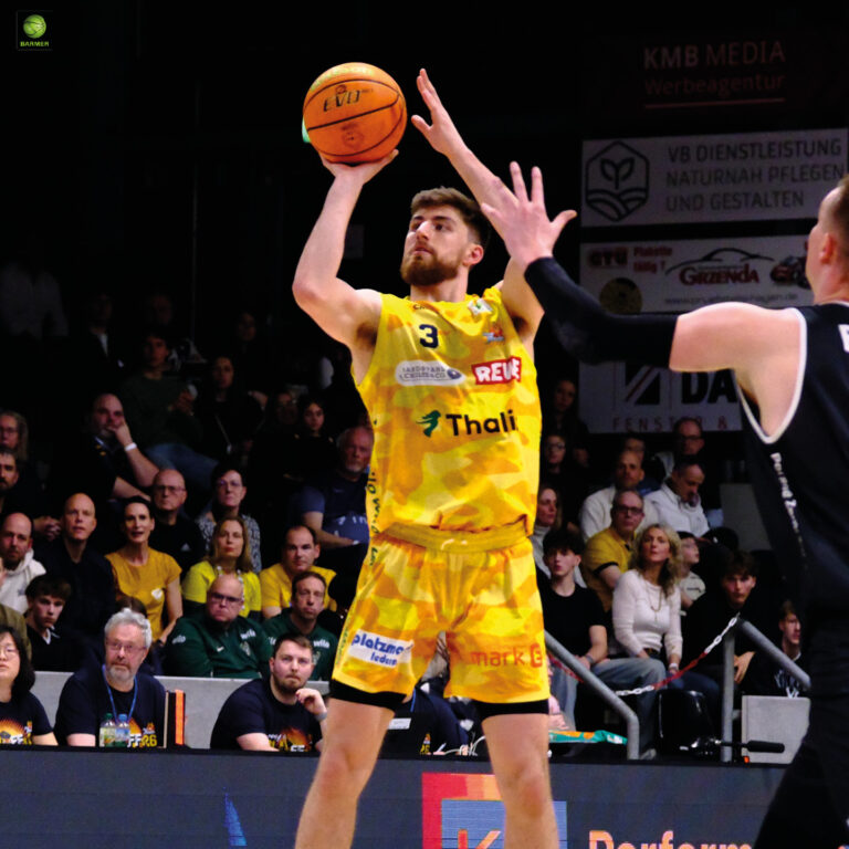 Ein Basketballspieler im gelben Trikot wirft konzentriert einen Ball während eines Spiels