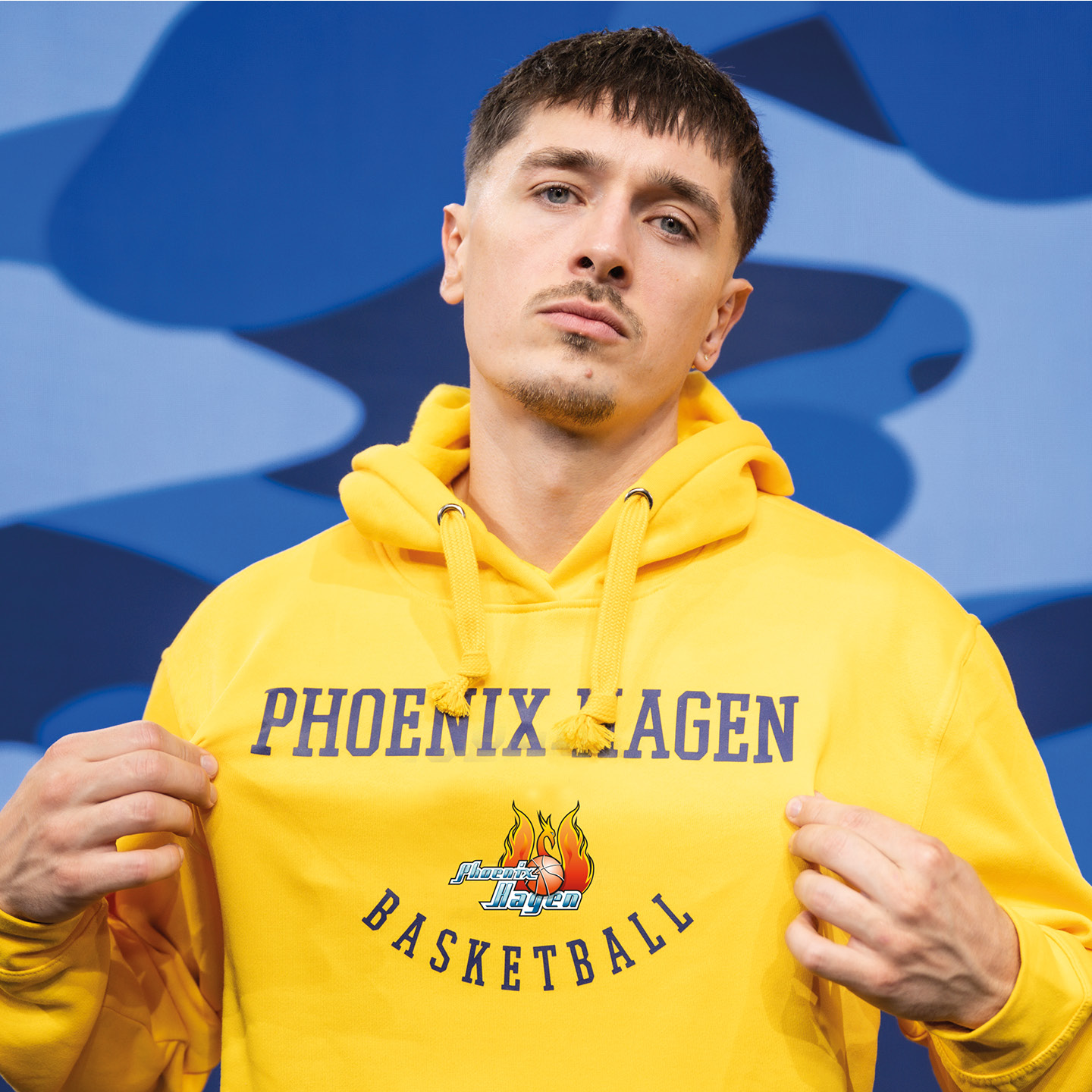 Ein junger Mann mit ernstem Blick trägt einen gelben Phoenix Hagen Basketball Hoodie