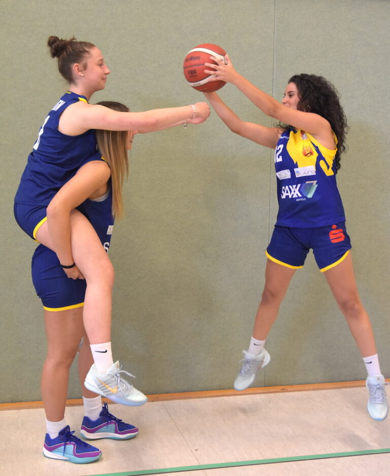 Drei Basketballspielerinnen in blauen Trikots, eine hebt lachend eine andere auf dem Rücken, während die dritte lächelnd den Ball hält