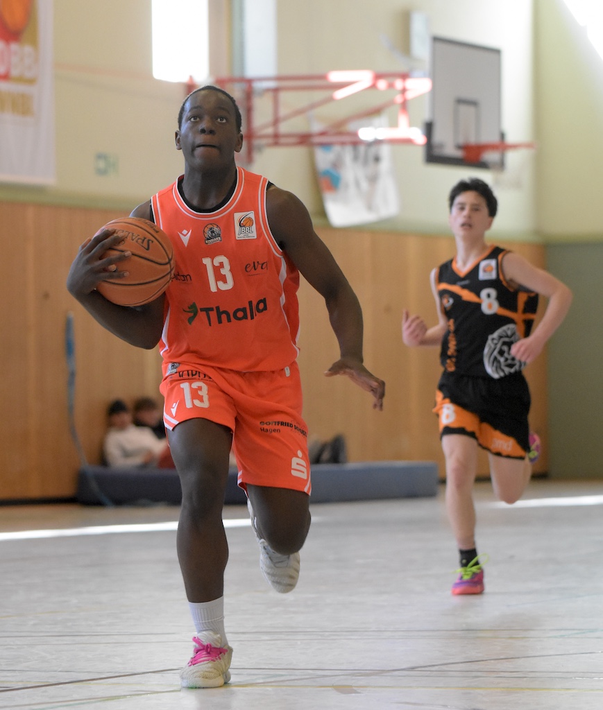 Ein junger Basketballspieler im orangefarbenen Trikot sprintet konzentriert aufs Korb zu, während ein Gegenspieler in schwarzem Trikot hinter ihm herläuft