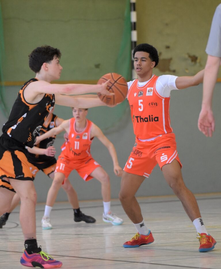 Drei junge Basketballspieler in orangefarbenen Trikots spielen konzentriert in der Halle