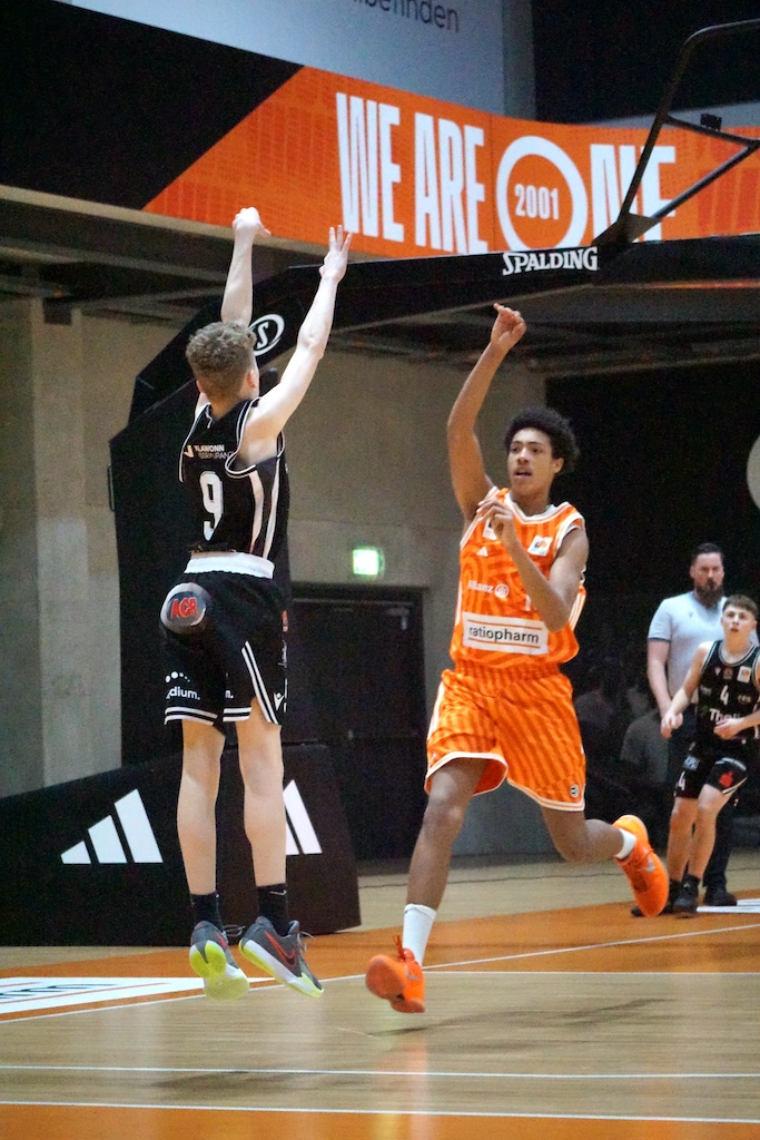 Zwei Basketballspieler im Spiel, einer in Schwarz springt zum Wurf, der andere in Orange verteidigt energisch