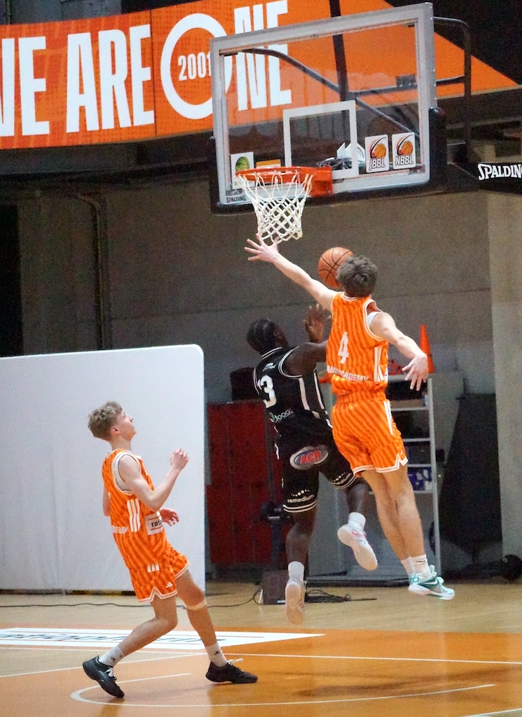 Zwei junge Basketballspieler in orangenen Trikots versuchen, gegen einen Gegner zu punkten