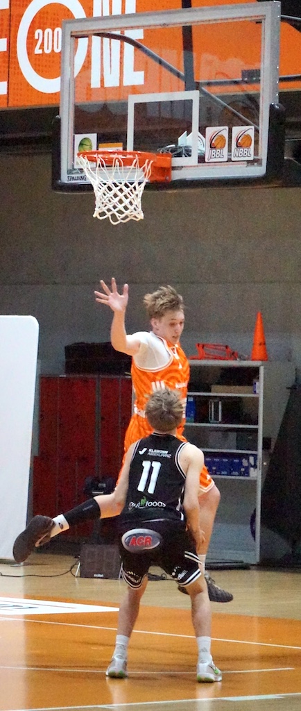 Zwei männliche Basketballspieler im Spiel, einer verteidigt dynamisch am Korb in Orange