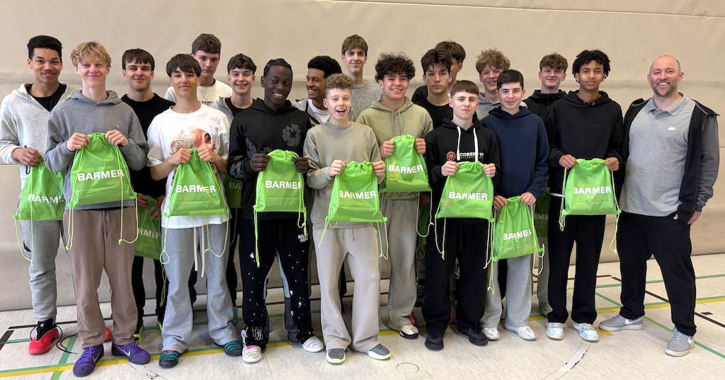 Eine Gruppe Jugendlicher und ein Erwachsener stehen fröhlich in einer Turnhalle und halten grüne Barmer Beutel