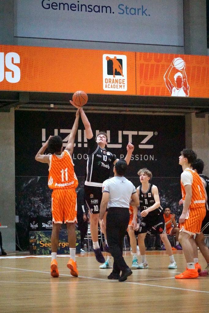 Zwei Basketballteams in Orange und Schwarz kämpfen konzentriert um den Ball beim Sprungball