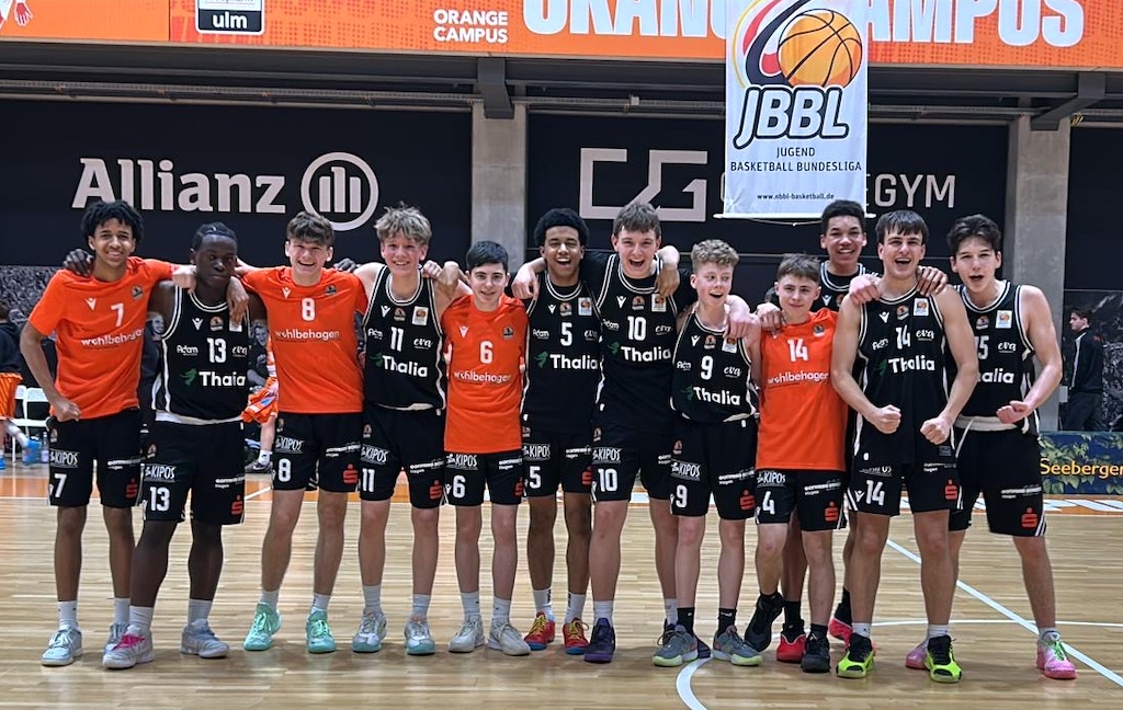 Eine Jugendbasketballmannschaft mit fröhlichen Gesichtern posiert in orangener und schwarzer Sportkleidung auf einem Hallenboden