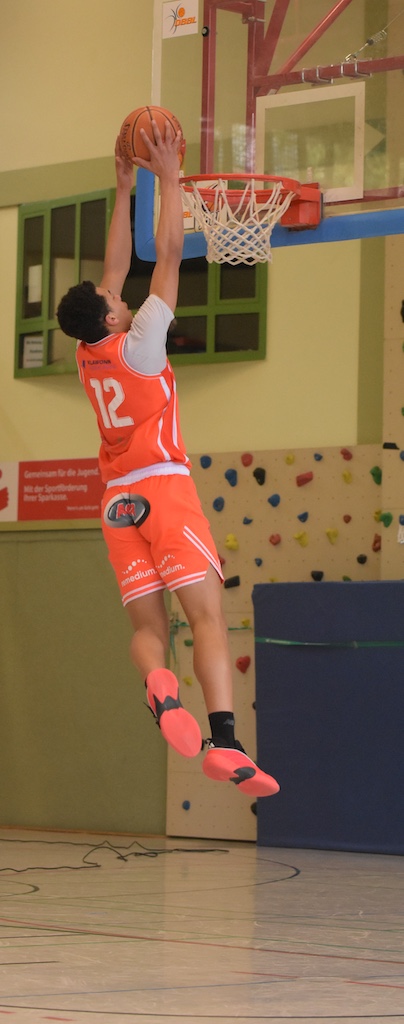 Ein junger Basketballspieler mit oranger Nummer 12 springt lächelnd zum Korb hoch