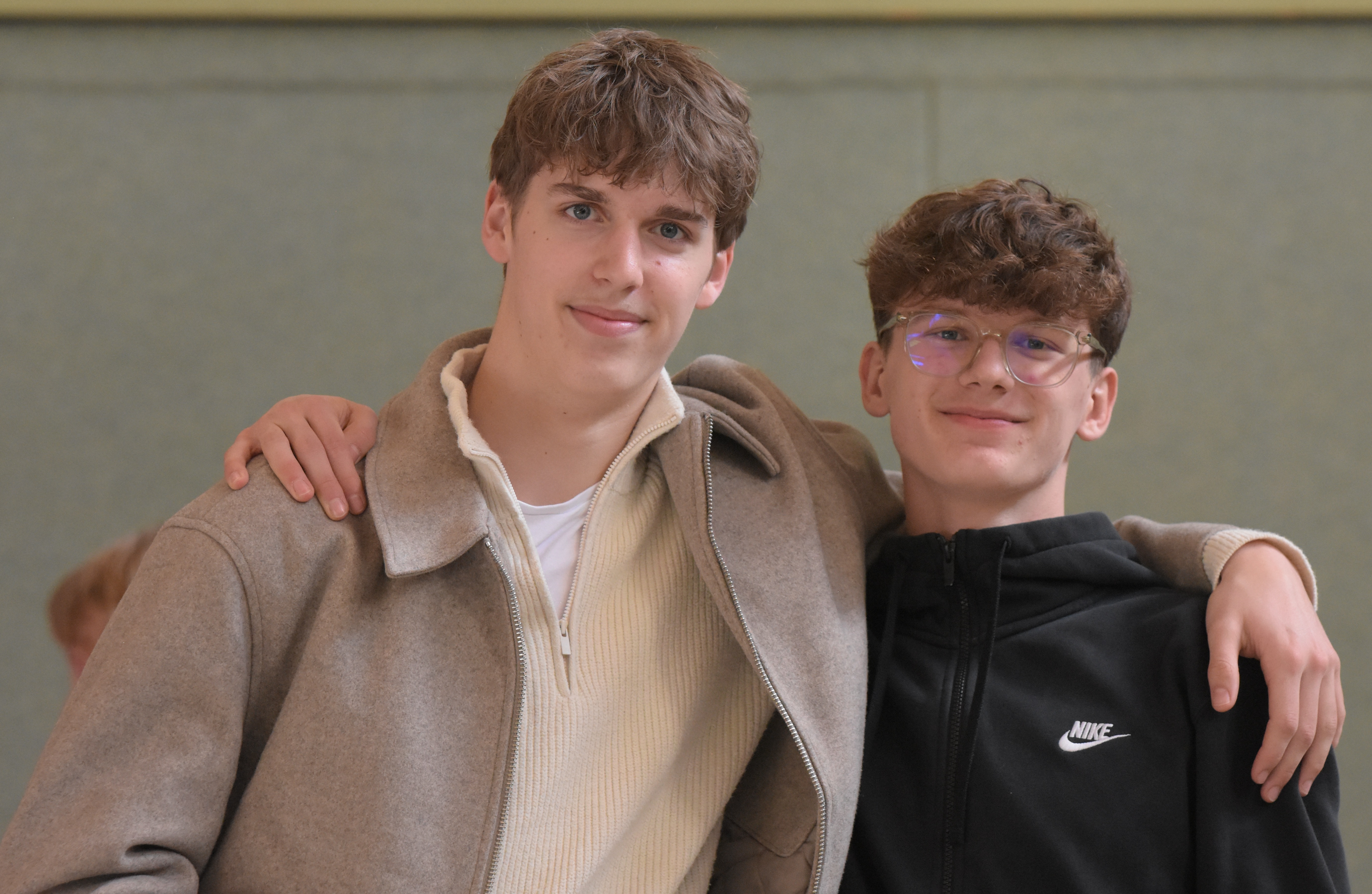 Zwei junge Männer lächeln freundlich in die Kamera, einer trägt eine Brille und Nike Jacke