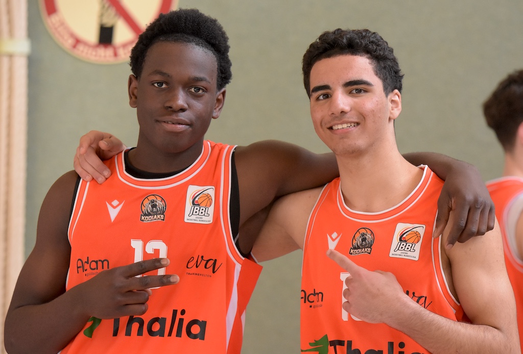 Zwei junge Basketballspieler in orangefarbenen Trikots lächeln freundlich in die Kamera