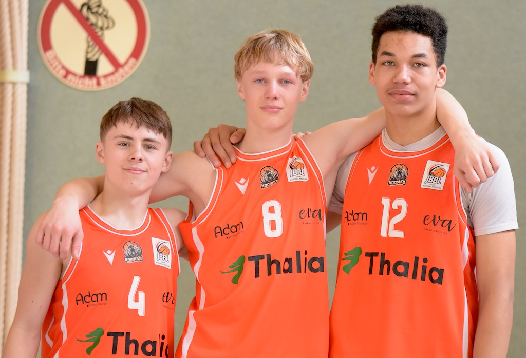 Drei junge Basketballspieler mit orangefarbenen Trikots lächeln und stehen Arm in Arm