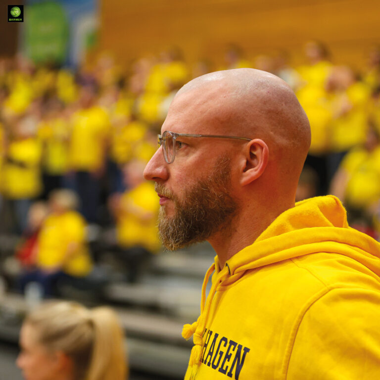 Ein glatzköpfiger Mann mit Bart blickt konzentriert im gelben Hoodie zur Seite