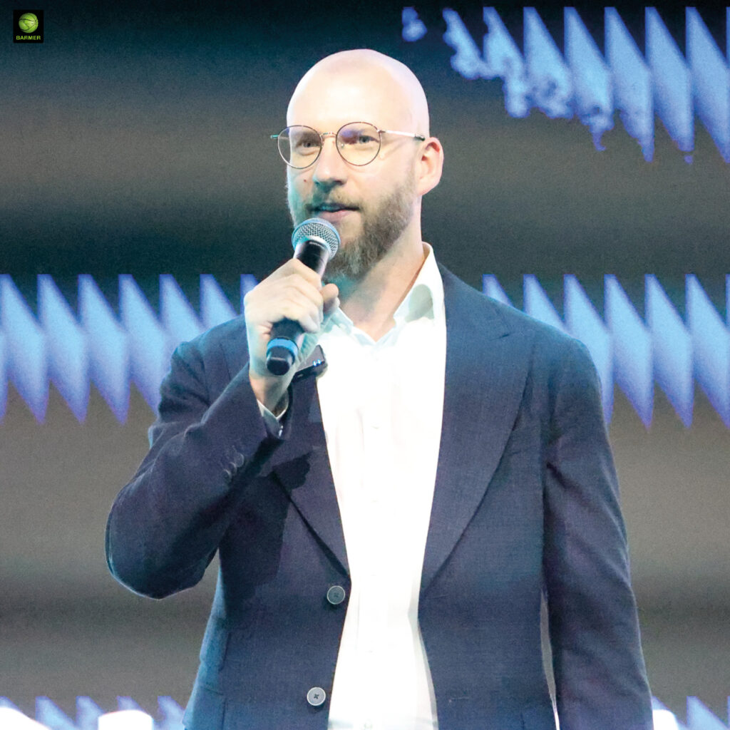 Ein glatzköpfiger Mann mit Brille hält ein Mikrofon und trägt einen dunkelblauen Anzug