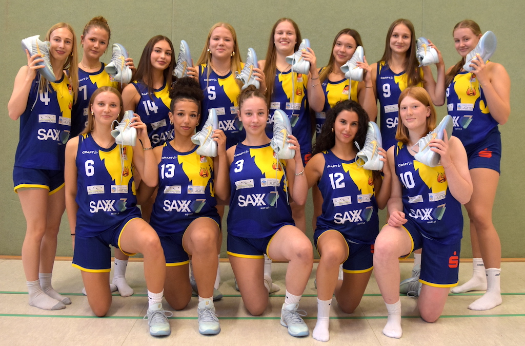 Eine Frauen-Volleyballmannschaft in blauen Trikots posiert lächelnd mit Sportschuhen