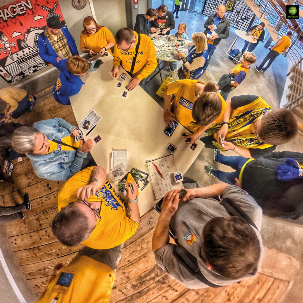 Menschen in gelber Kleidung tauschen fröhlich Sammelbilder an einem weißen Tisch