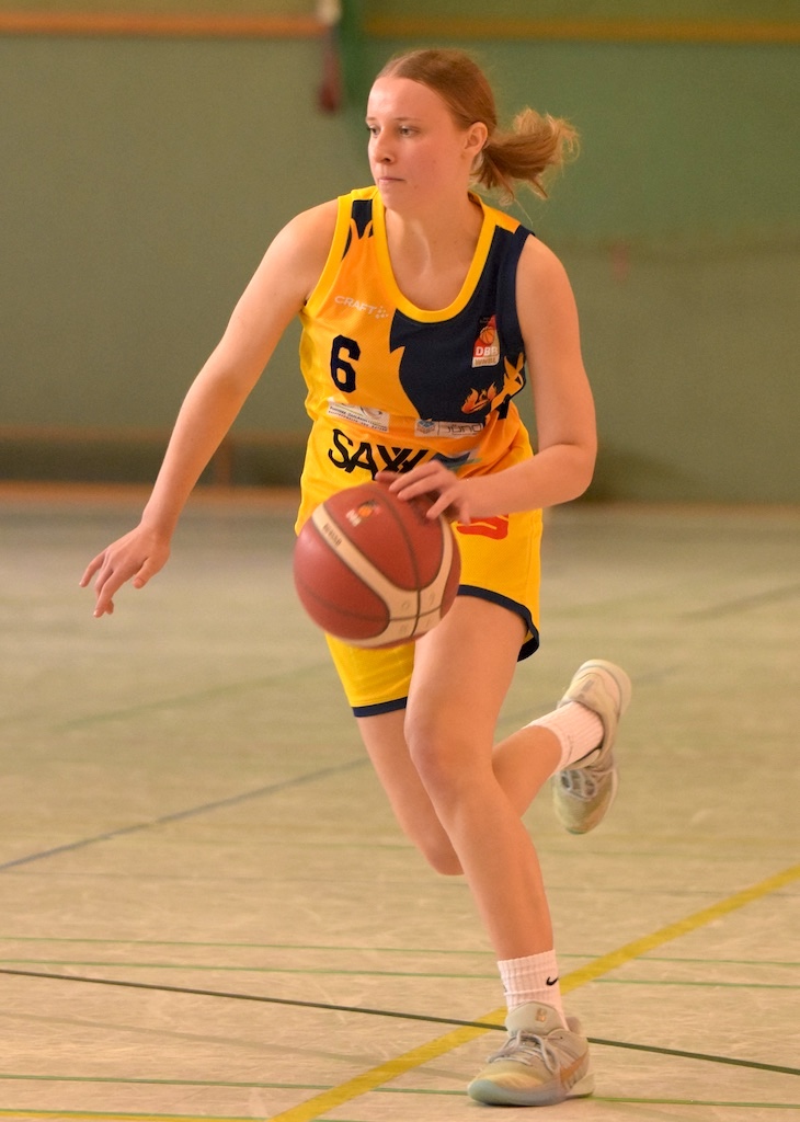 Eine junge Frau im gelben Basketballtrikot dribbelt konzentriert den Ball auf dem Spielfeld