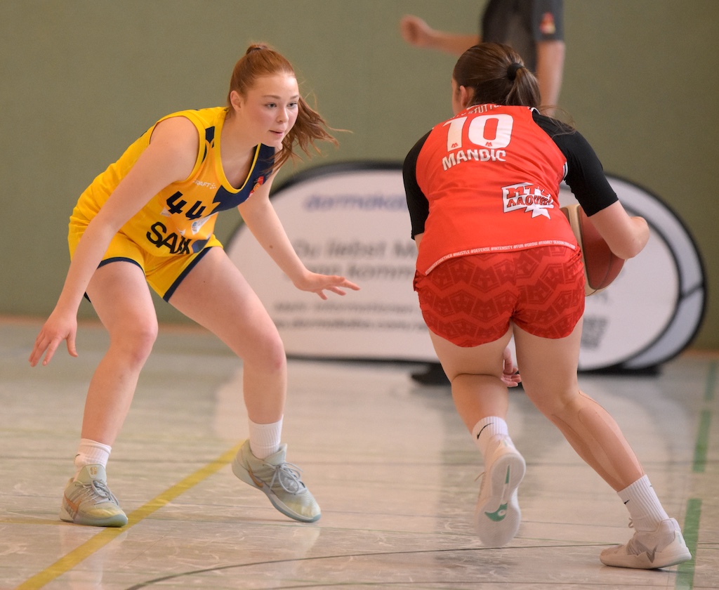Zwei Basketballspielerinnen im gelben und roten Trikot stehen sich konzentriert gegenüber