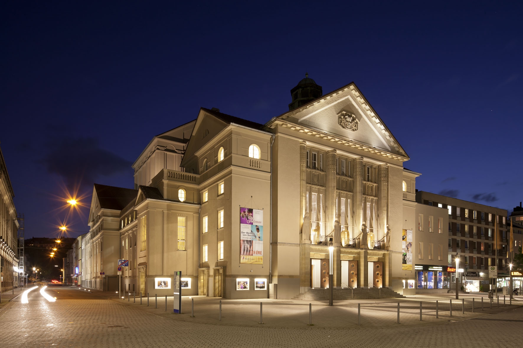 Das Bild zeigt ein beleuchtetes, historisches Theatergebäude bei Nacht in hellen Farben