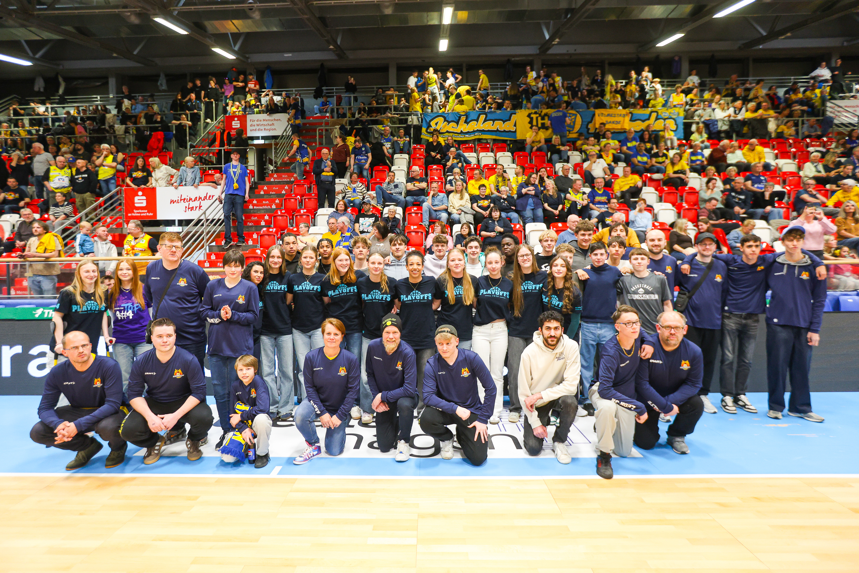 Mehrere Personen stehen und sitzen lachend in einer Sporthalle vor vollen Tribünen