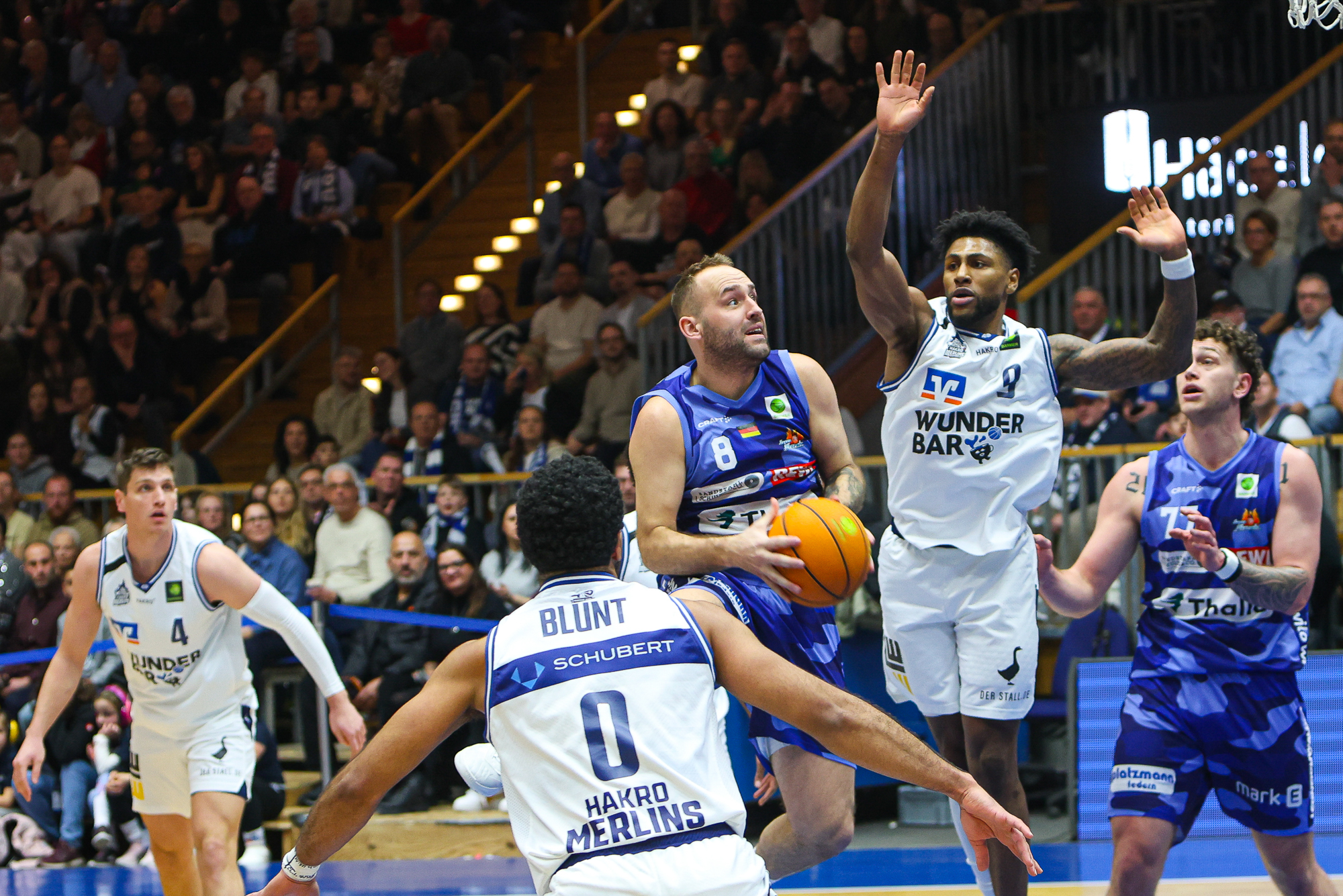 Ein Basketballspieler im blauen Trikot springt mit entschlossenem Gesichtsausdruck zum Korb, umgeben von Gegnern in weißen Trikots in einer vollen Halle