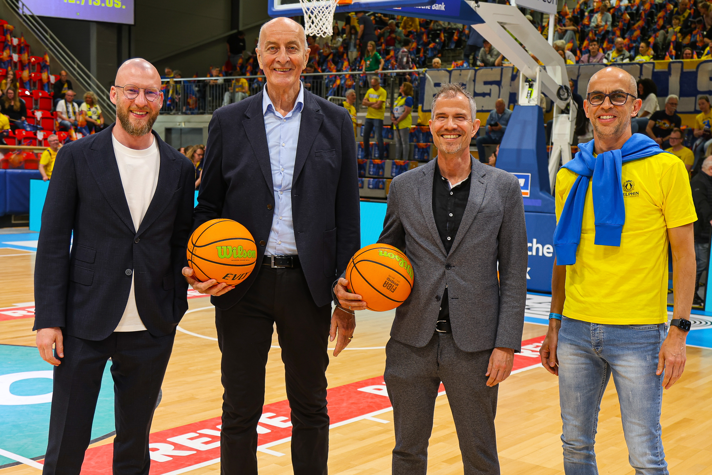Vier Männer stehen lächelnd auf einem Basketballfeld, zwei halten Basketbälle, einer trägt ein gelbes T-Shirt und blaue Pullover über den Schultern