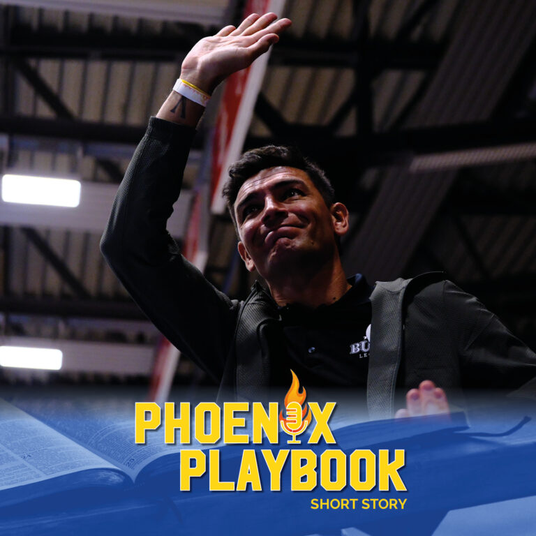 Ein dunkelhaariger Mann lächelt freundlich und winkt unter Hallenbeleuchtung