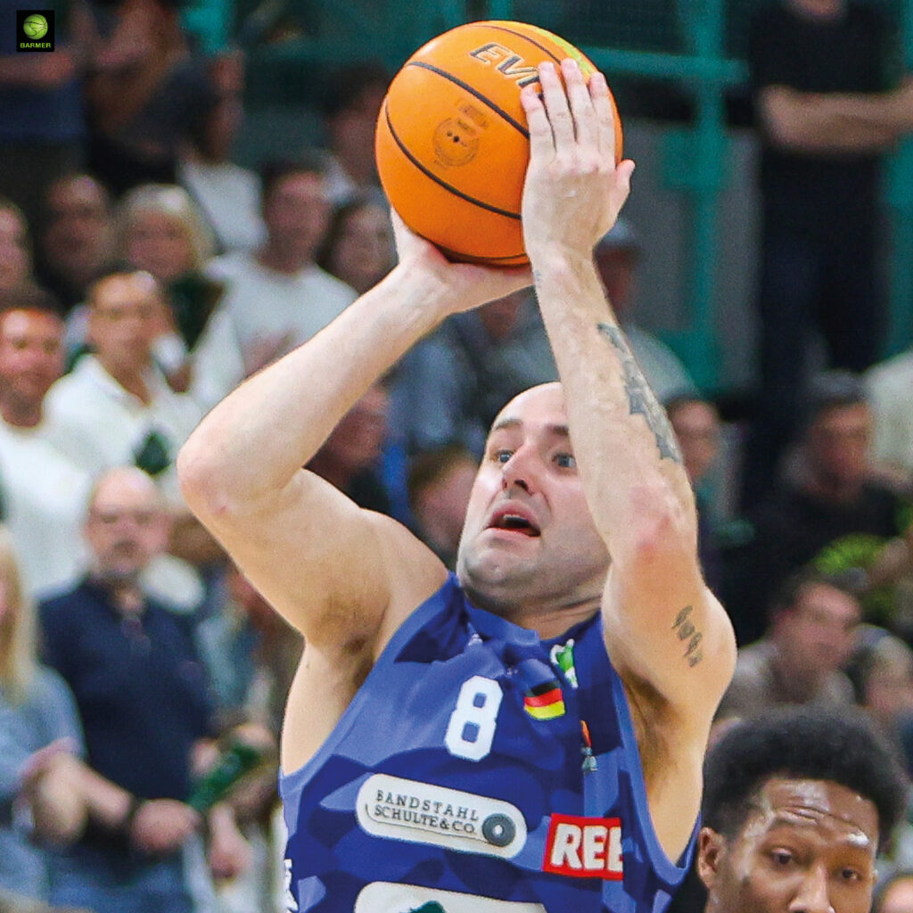 Ein Basketballspieler mit fokussiertem Blick wirft den orangen Ball im blauen Trikot
