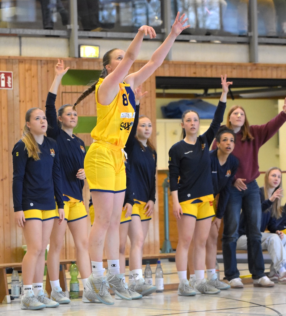 Eine junge Basketballspielerin in gelb wirft konzentriert den Ball, Team jubelt im Hintergrund