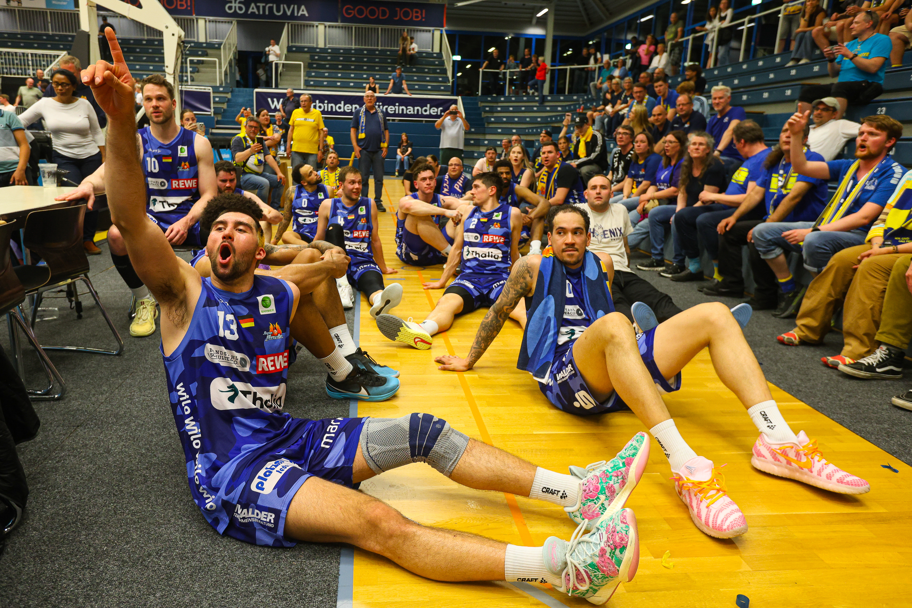 Mehrere Basketballspieler im blauen Trikot jubeln lachend am Spielfeldrand auf dem Boden
