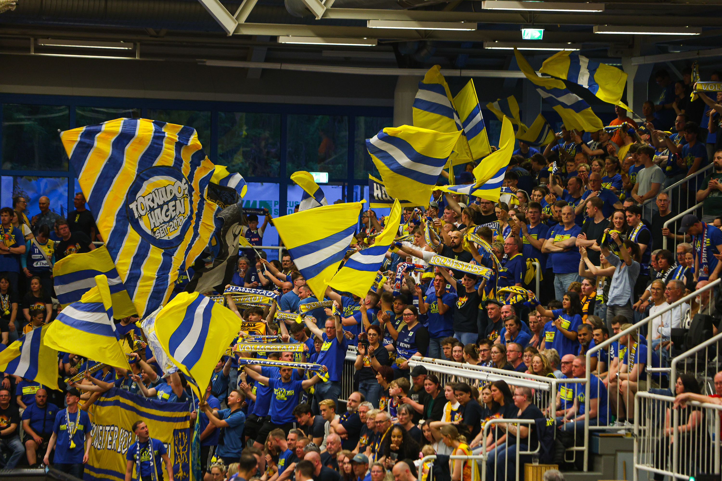Viele jubelnde Fans mit gelben und blauen Fahnen bei einem Sportevent in der Halle