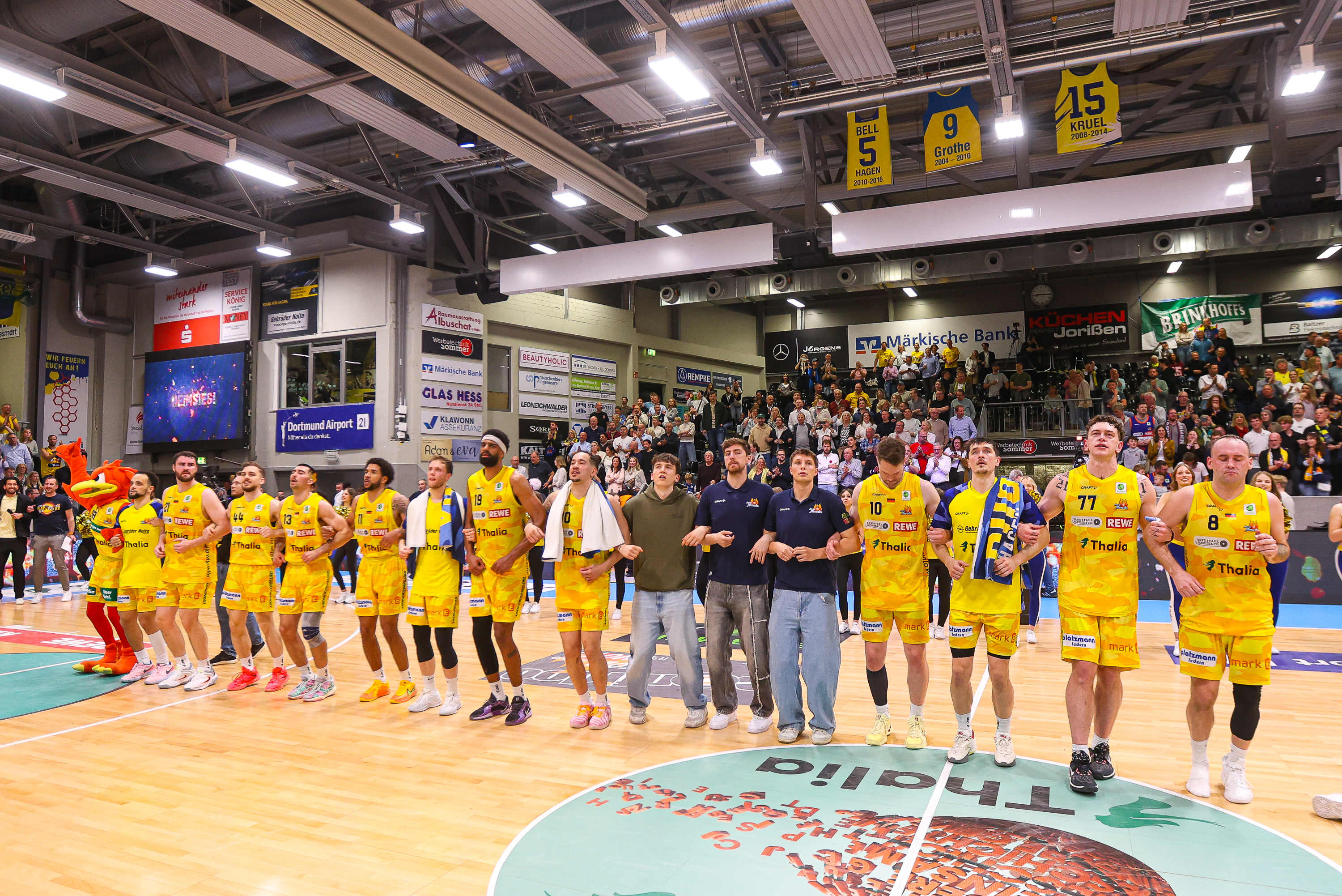 Mehrere Basketballspieler im gelben Trikot lächeln gemeinsam mit Fans auf dem Spielfeld