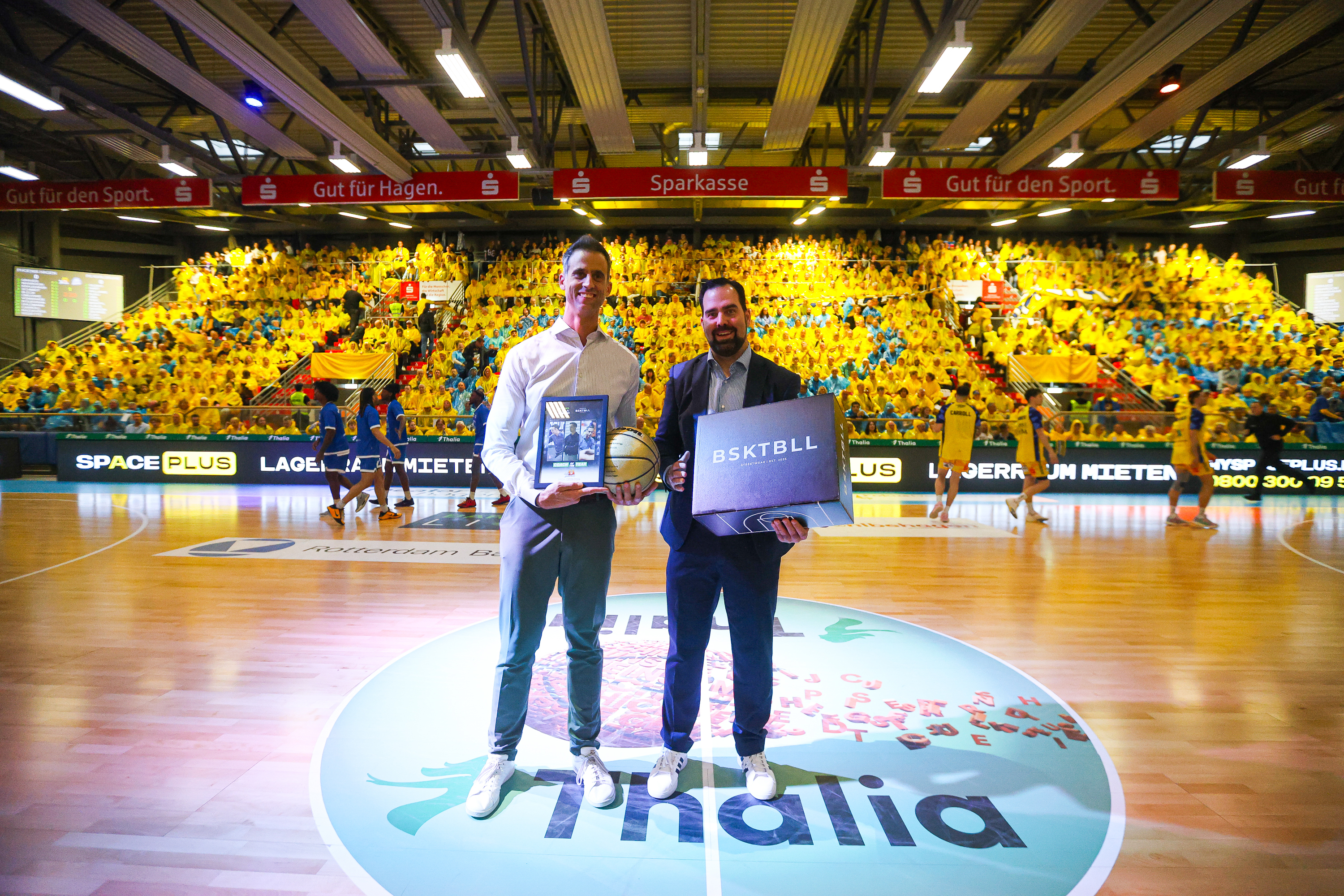 Zwei Männer stehen lächelnd auf einem Basketballfeld mit Geschenken in den Händen