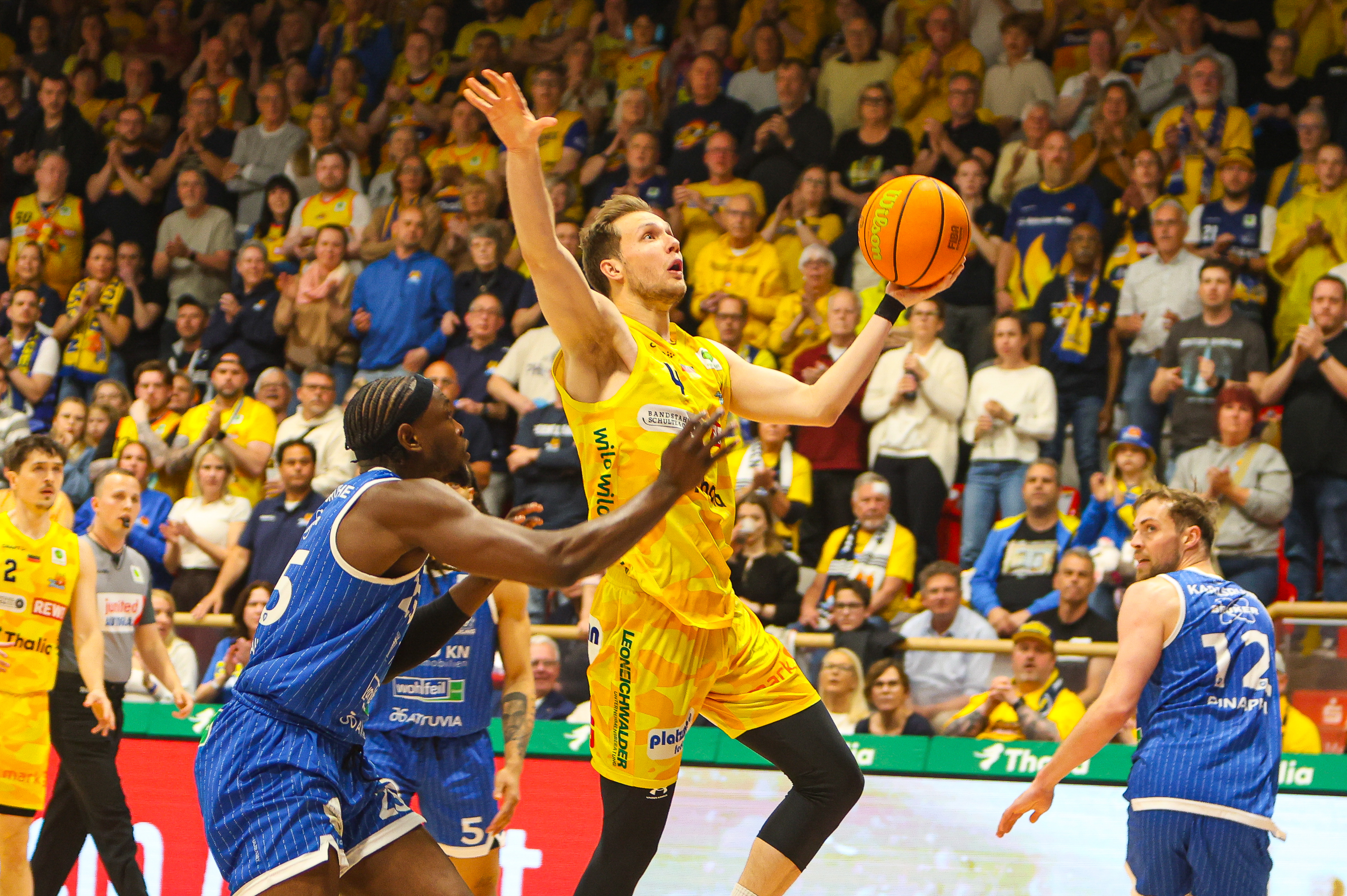 Ein Basketballspieler im gelben Trikot springt entschlossen zum Korb vor Publikum