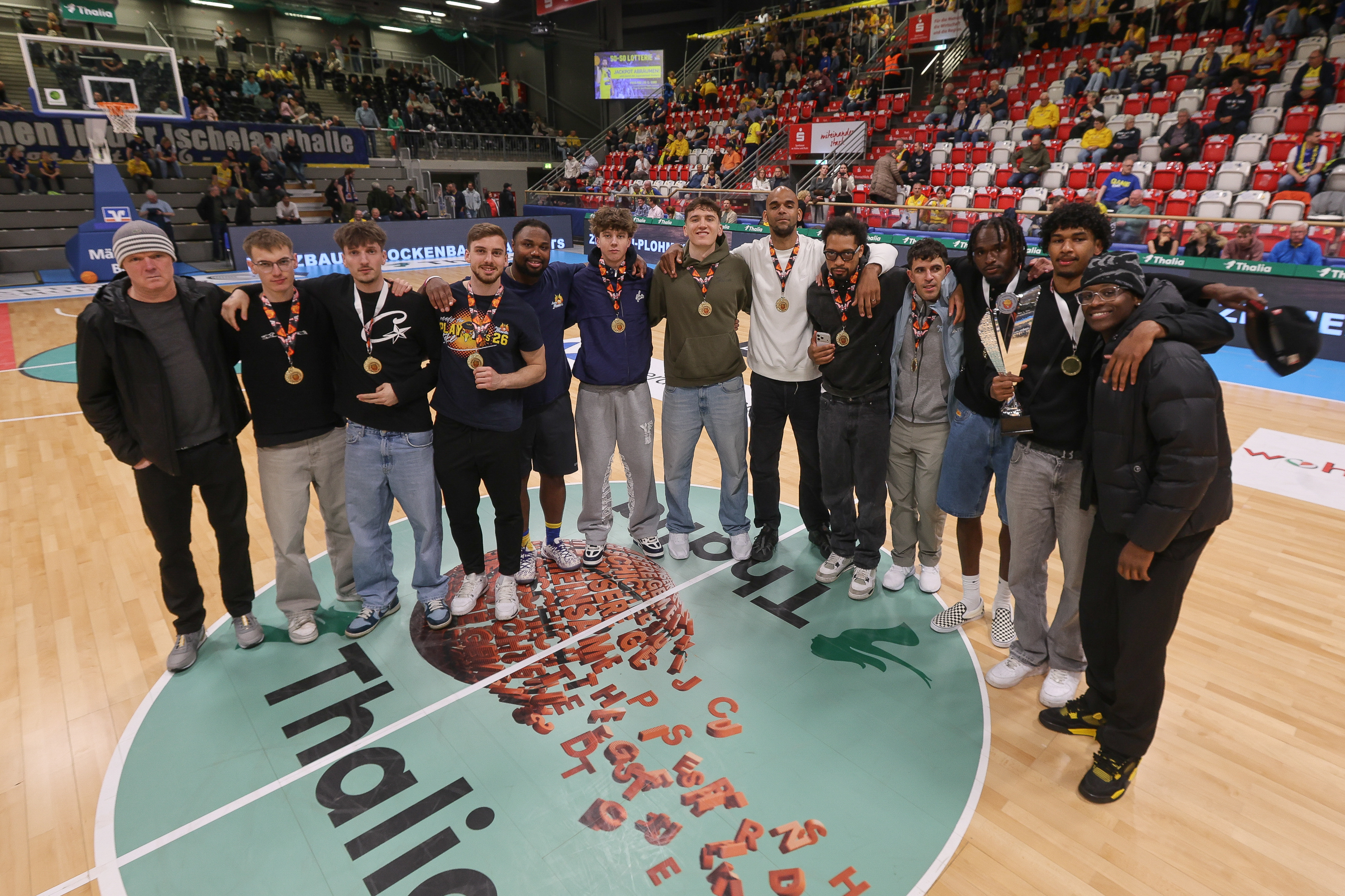 Eine Gruppe junger Männer steht lachend auf einem Basketballfeld mit Medaillen um den Hals