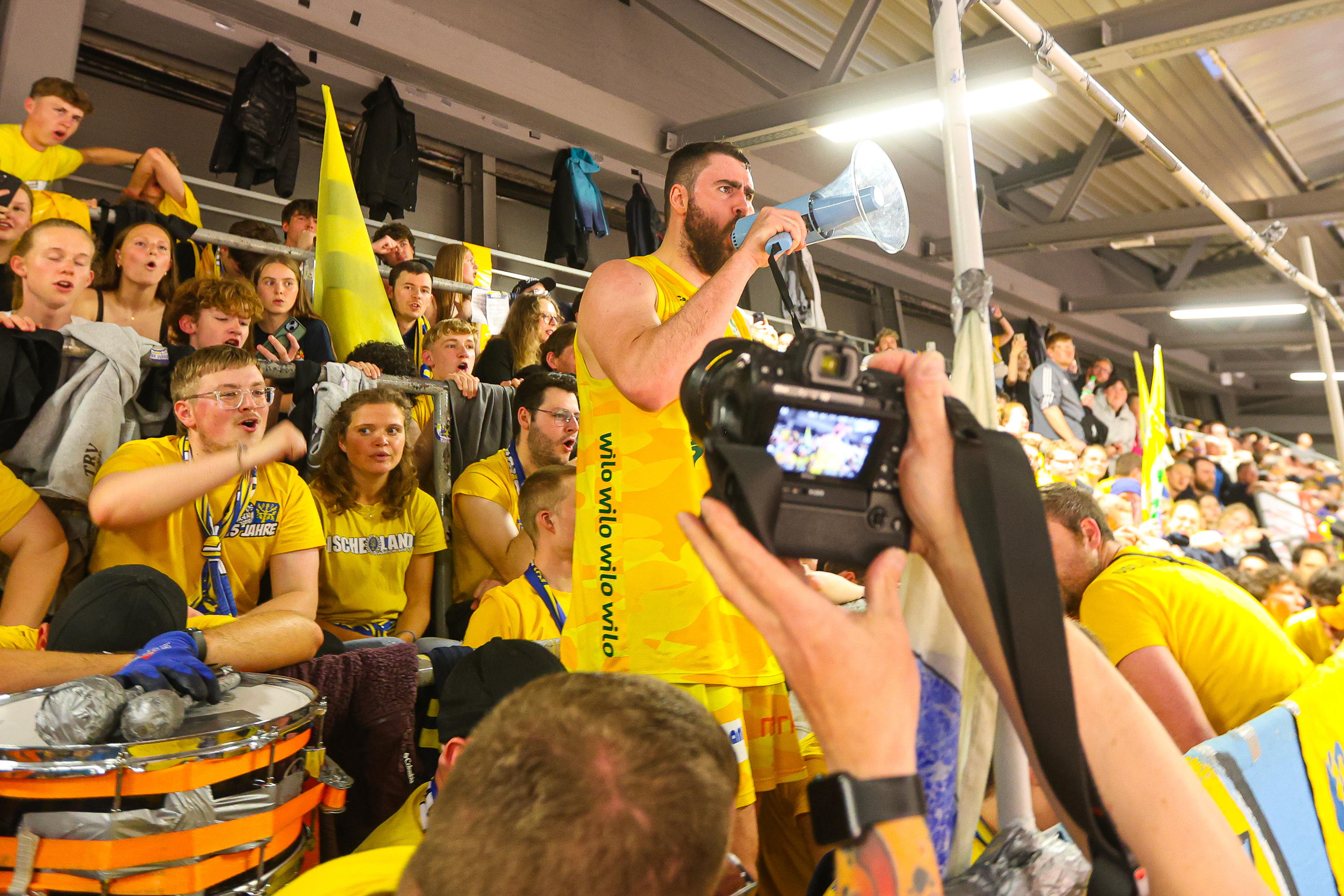 Ein Mann im gelben Trikot ruft mit Megafon vor begeisterten Fans im Stadion